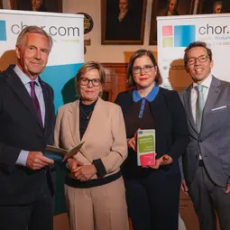 Zwei Männer und zwei Frauen in schicker Kleidung stehen vor einem Schild auf dem chor.com steht, der Name einer Musikveranstaltung